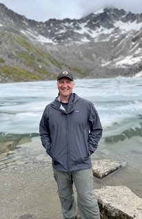 A person wearing a jacket stands in front of a lake and snow-covered mountains