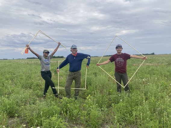 Three smiling people pose in a field, holding one meter quadrats 